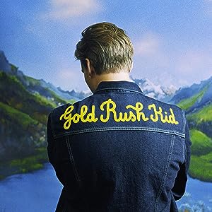 A person with 'GOLD RUSH KID' text on their denim jacket standing in front of a landscape painting.