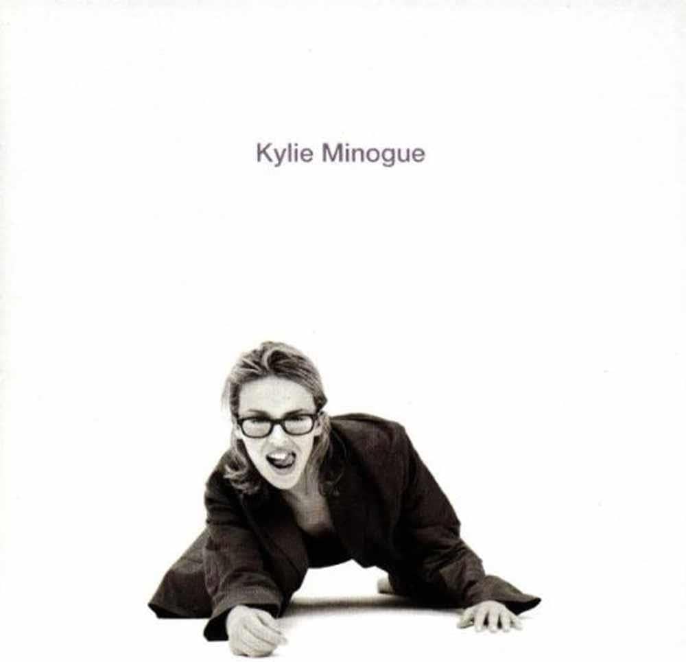 Album cover for Kylie Minogue, featuring a black and white photograph of the artist crawling on the floor with her mouth open, wearing glasses and a dark jacket, against a solid white background with her name printed at the top.