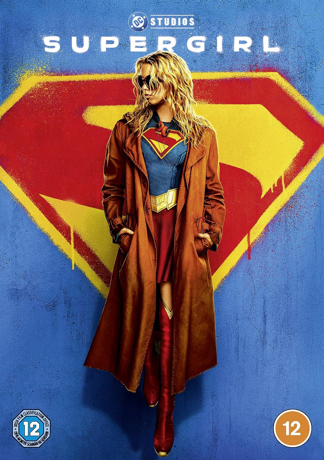 DC Studios Supergirl poster featuring a woman in a brown trench coat over a blue, red, and yellow costume, standing in front of a large red and yellow Superman-style shield on a blue background.