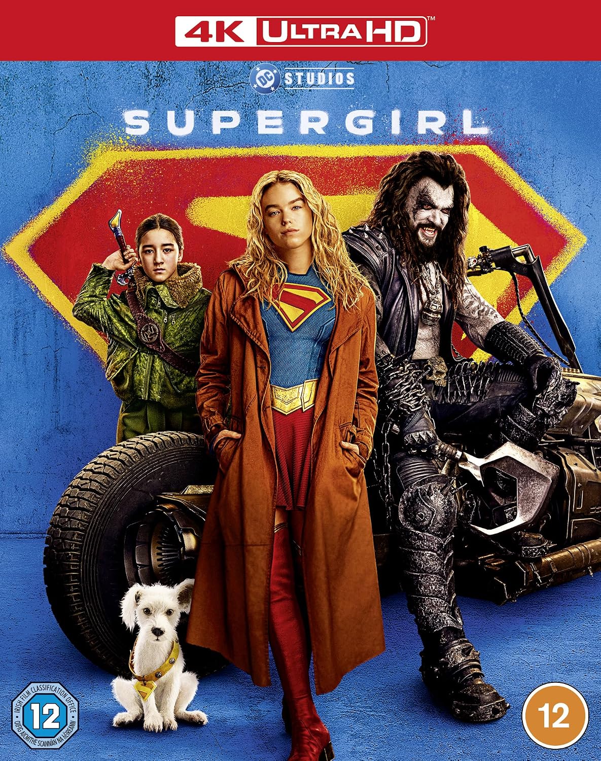 4K Ultra HD movie poster for 'Supergirl' featuring a young woman in a red and blue superhero costume standing in front of a large red and yellow shield, with another person in green behind her, a masked figure seated on a motorcycle to the right, and a small white dog at the bottom left. The top banner reads '4K ULTRA HD' and the title 'SUPERGIRL' is centered.