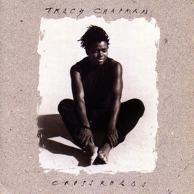 Album cover for Tracy Chapman's 'Crossroads', featuring a black-and-white portrait of the artist sitting cross-legged against a textured beige background with handwritten text.