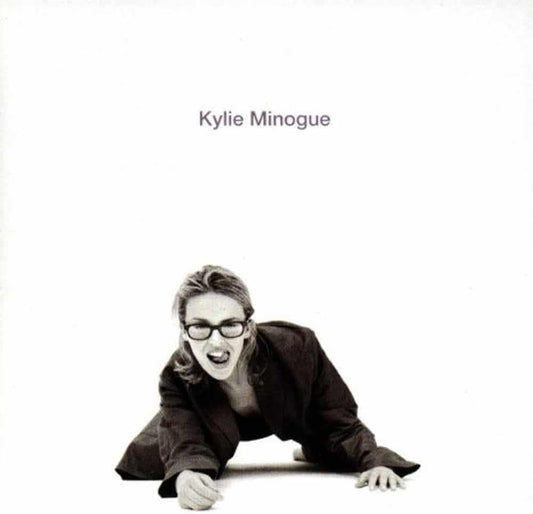 Album cover for Kylie Minogue, featuring a black and white photograph of the artist crawling on the floor with her mouth open, wearing glasses and a dark jacket, against a solid white background with her name printed at the top.
