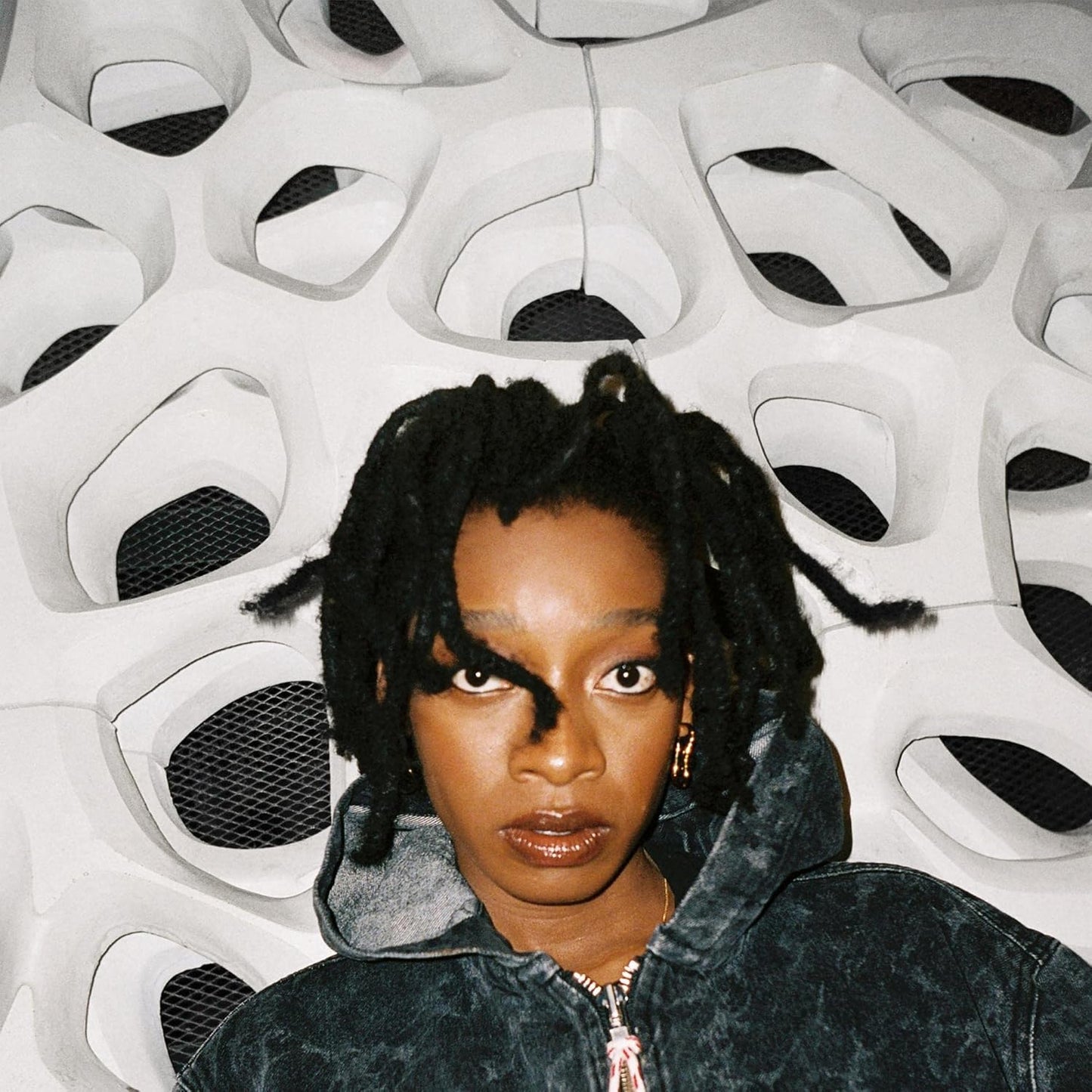 Person with braided hair wearing a dark jacket in front of a geometric white wall.