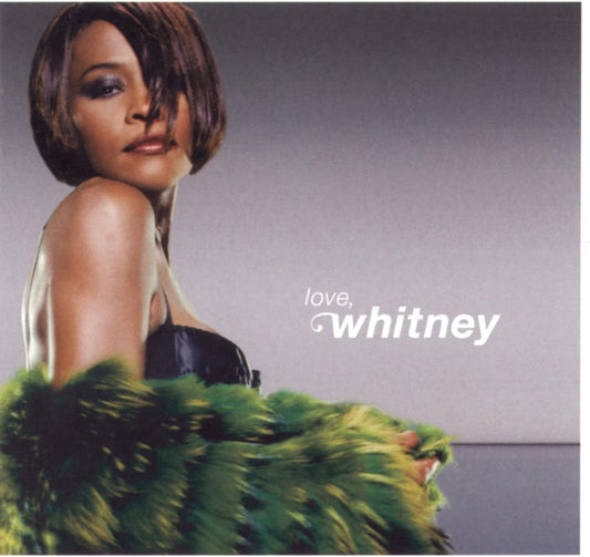 Album cover for Whitney 'Love, Whitney' featuring a woman with short dark hair looking over her shoulder, wearing a black strapless top and a voluminous green feathered skirt, with the title text on a gray gradient background.