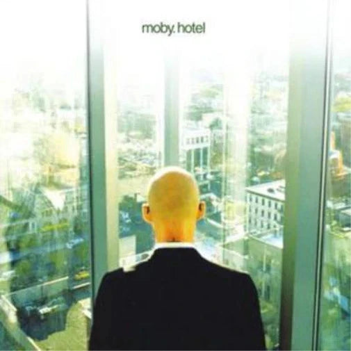 Album cover art for 'moby, hotel' featuring a bald man in a black suit seen from behind, looking out of a tall window over a cityscape. The title 'moby, hotel' is printed at the top in a small green font.