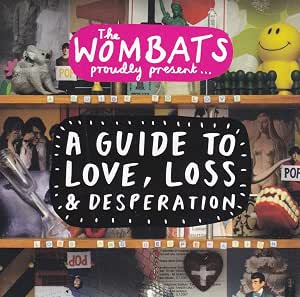Album cover for 'A Guide to Love, Loss & Desperation' by 'The Wombats', featuring a collage of various objects on shelves, including a yellow smiley face, a heart-shaped heart, and a small dog.