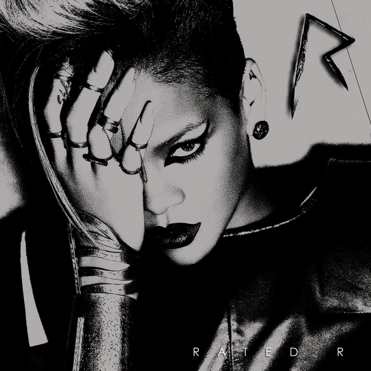 Black and white close-up portrait of a person with dramatic eye makeup, dark lipstick, rings, and a high-top hairstyle, with a dark jacket and a stylized letter graphic in the background.