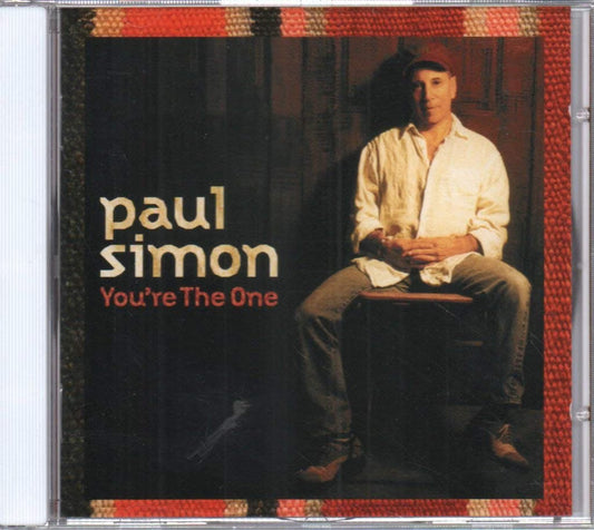 CD cover for Paul Simon's album 'You're The One' in a jewel case. The cover features a dark background with Paul Simon seated on a stool, wearing a white shirt and jeans, with the text 'paul simon' and 'You're The One' in beige and red lettering.